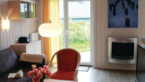 Un salon comprenant un canapé, un fauteuil rouge, une table avec un livre et des fleurs, une lampe, une cheminée et une vue sur la verdure à travers une porte vitrée.