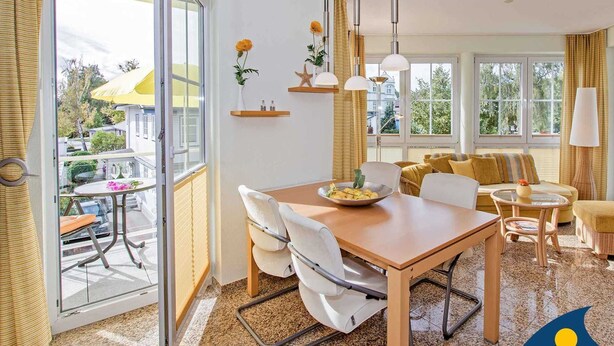 Un salon moderne avec une table à manger en bois, des chaises blanches, un canapé et une grande fenêtre donnant sur l’extérieur.