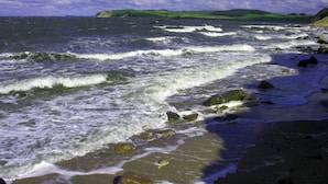 En strand med vågor, klippor och ett avlägset, grönt kullområde.