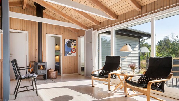 Una habitación acogedora con techo de madera, una chimenea, dos sillones negros con cojines estampados, una mesita con adornos y un ventanal con vistas al exterior.