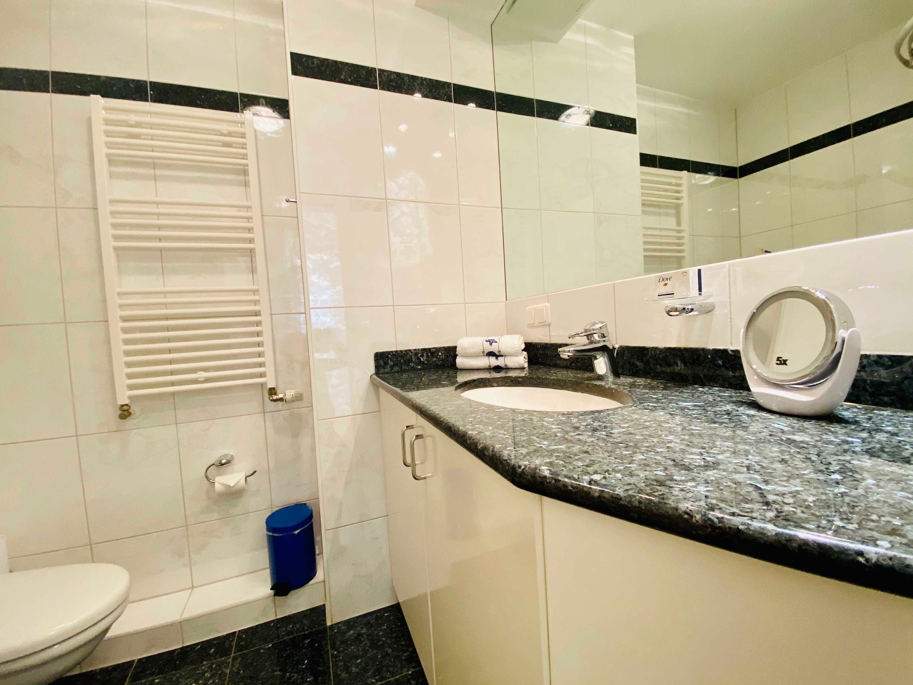 A bathroom with a toilet, sink, mirror, towel dispenser, and a blue trash bin.