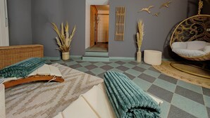 A room with a checkered floor, a bed with a white and teal blanket, a wicker chair with a white cushion, and a hanging chair with a patterned design. - Residence Bleichröder, App. 27 - Residence Bleichröder Villa Rosengarten (Heringsdorf)