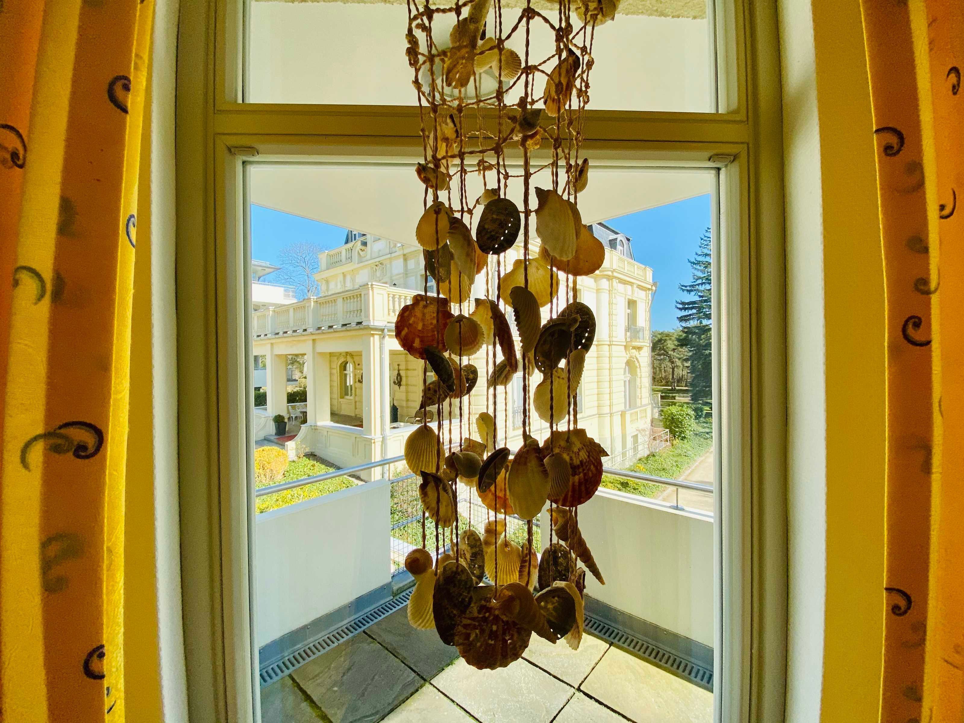 Blick durch ein Fenster mit einer hängenden Muscheldekoration, ein weißes Gebäude im klassischen Stil und ein klarer, blauer Himmel.