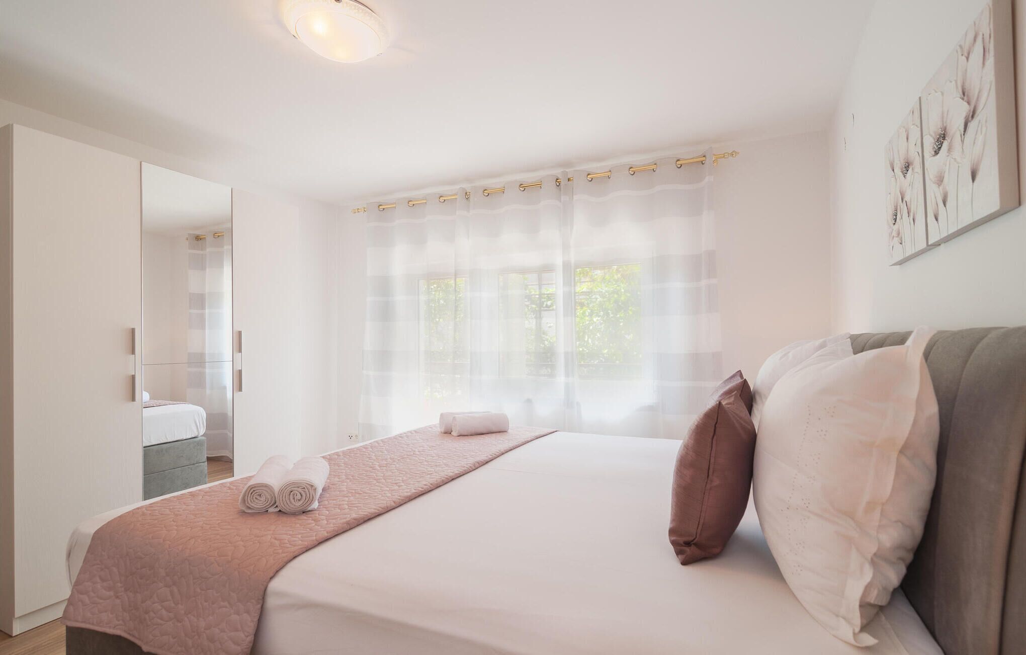 A modern bedroom with a large bed, a bedside table, a mirror, and a window with sheer curtains.
