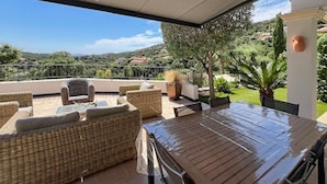 Un patio exterior con mobiliario de mimbre, una mesa de vidrio y vistas a un paisaje verde.