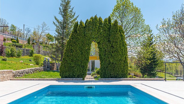 A swimming pool with a clear blue water, surrounded by a stone wall and a well-maintained garden with various plants and a tall, conical hedge. - Luxurious home in Kijevo (Kijevo)