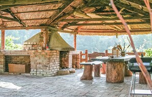 Un espace cuisine extérieur doté d’un four en briques, de tables en bois et d’une structure à toit de chaume.