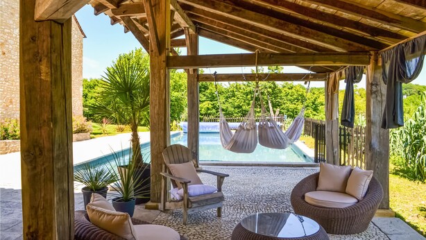Un espacio de estar al aire libre cubierto, con piscina, mobiliario de madera y plantas en macetas.