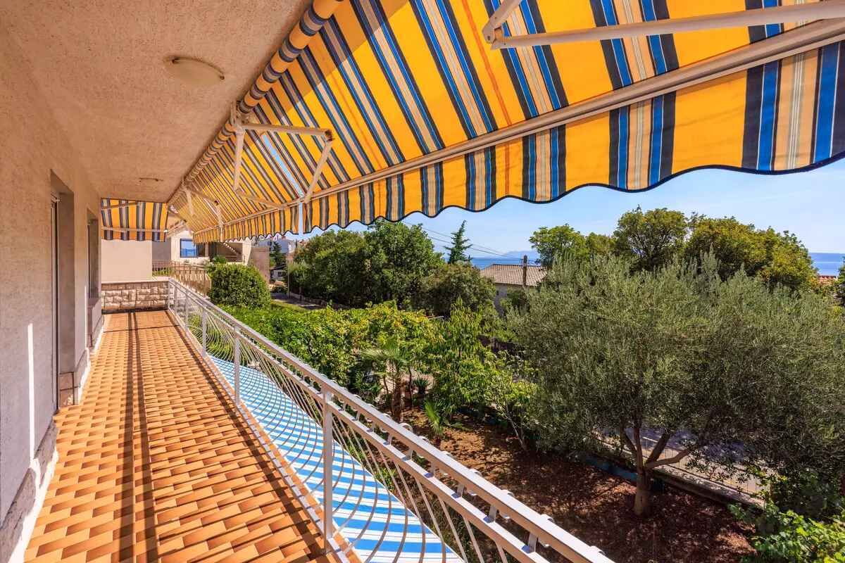 A balcony with a striped awning, a tiled floor, and a view of greenery and the sea.