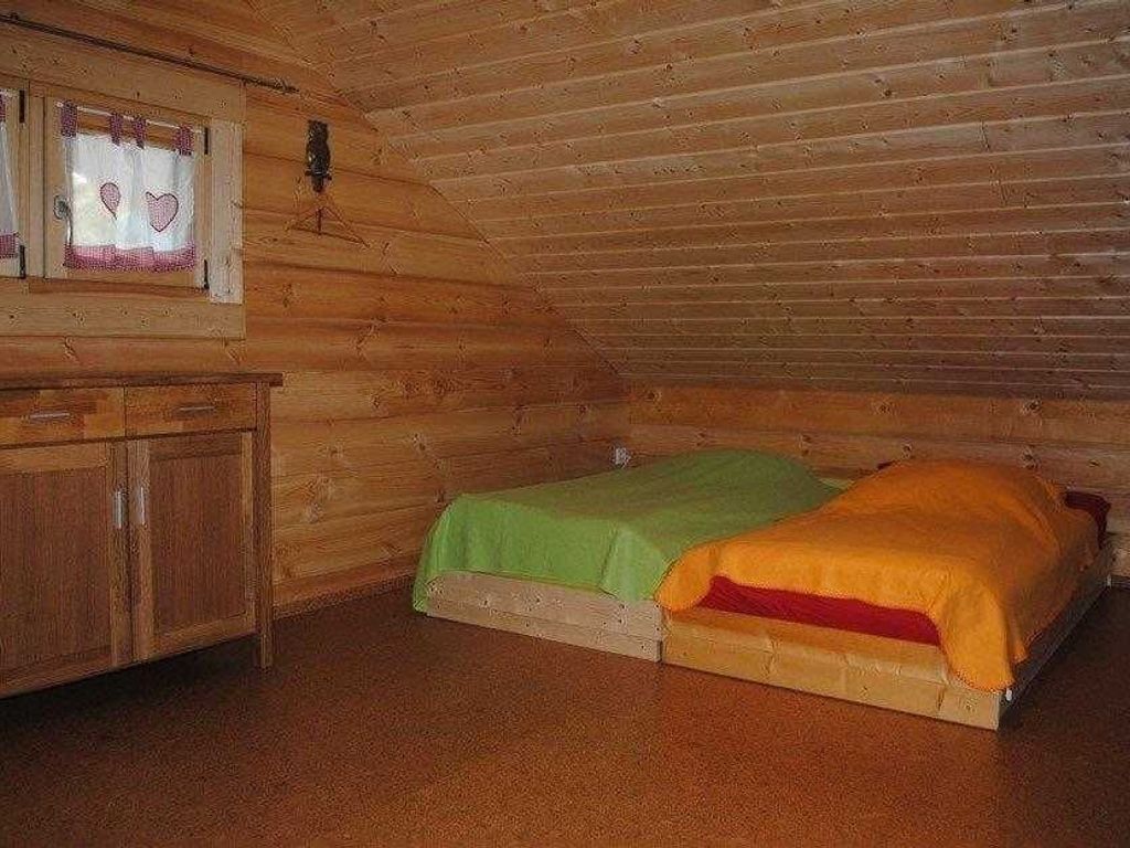 A small, cozy room with a wooden ceiling and walls, featuring a single bed with green and orange bedding, a wooden dresser, and a window with heart-shaped decorations.