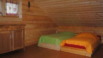A small, cozy room with a wooden ceiling and walls, featuring a single bed with green and orange bedding, a wooden dresser, and a window with heart-shaped decorations.