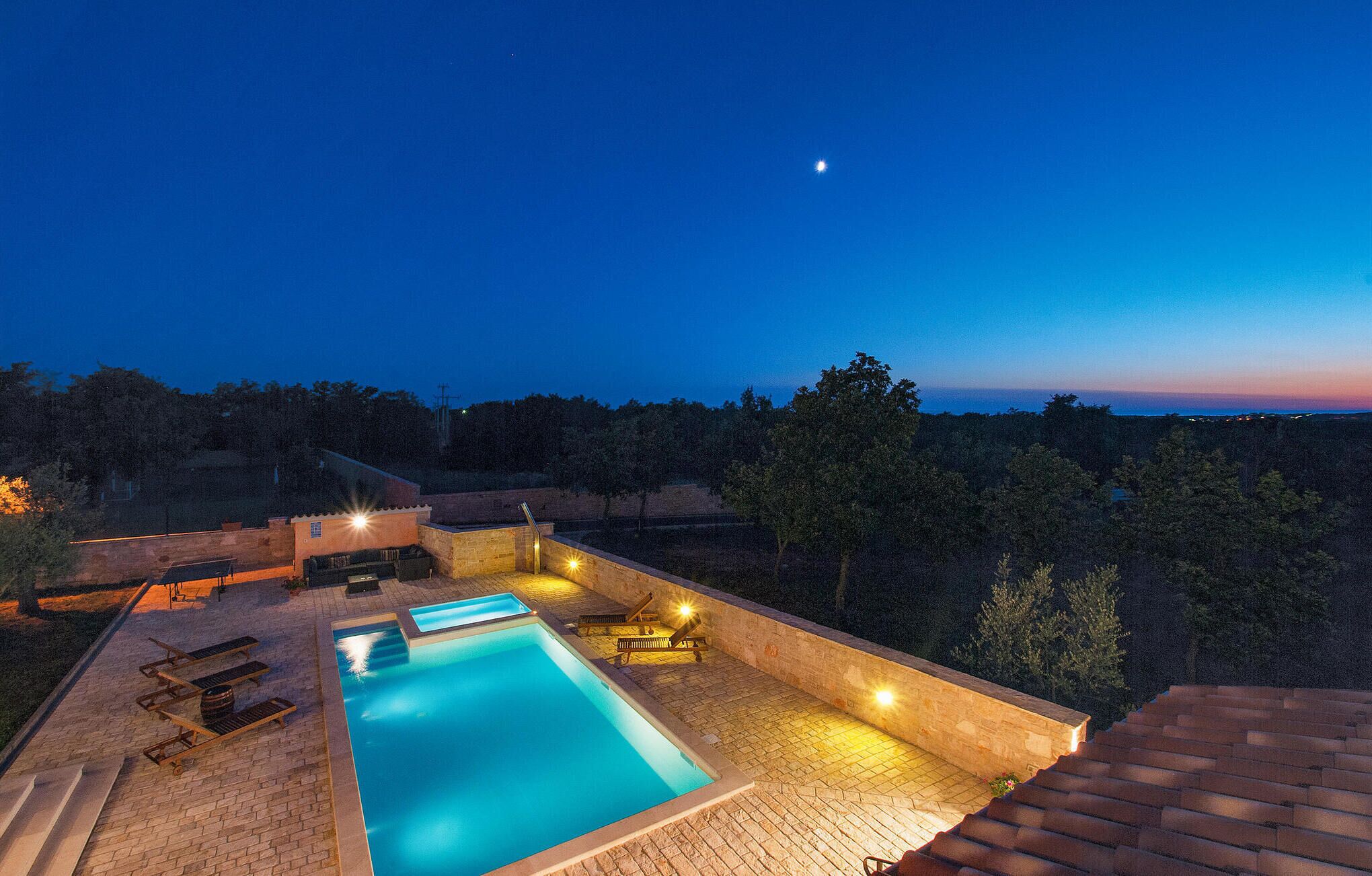 Une piscine offrant une vue dégagée sur le ciel nocturne, entourée d’une terrasse en pierre et de chaises longues.