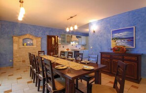 A dining area with a long wooden table set for six, flanked by chairs. A stone fireplace with a small shelf above it is visible. The room has blue walls and a large painting of a coastal scene. - Beautiful home in Vodnjan (Vodnjan)