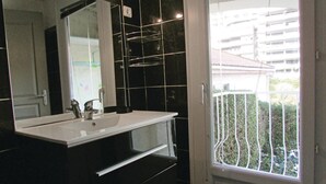 A modern bathroom with a white sink, black tiles, and a large mirror. There is a window with a metal railing and a view of buildings and greenery outside. - 1 bedroom nice apartment in Bastia (Bastia)