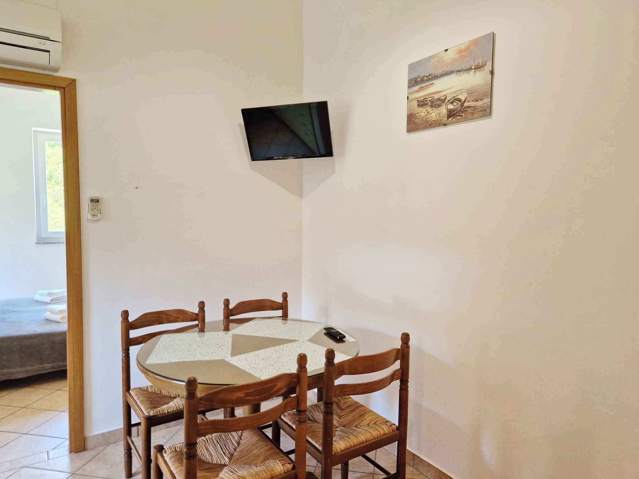 A small dining area with a round table and four wooden chairs, a television mounted on the wall, and a painting above the table.
