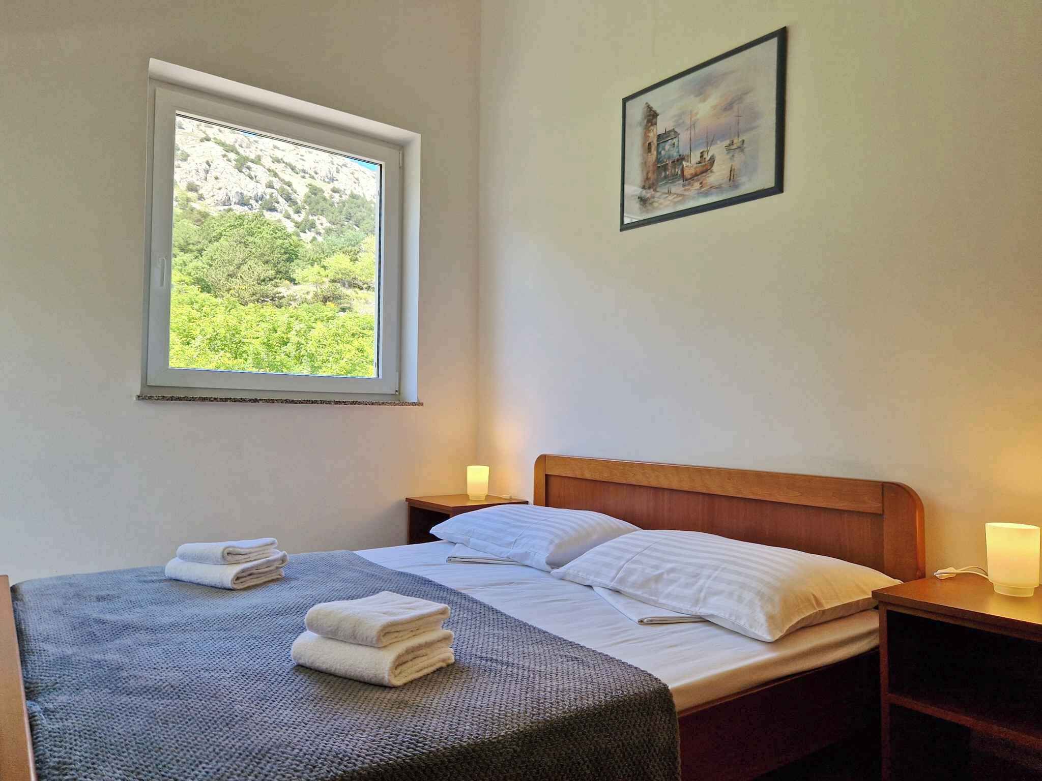A bedroom with a bed, a window with a view of greenery, a painting on the wall, and a bedside table with a lamp.