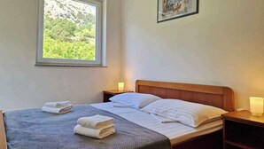 A bedroom with a bed, a window with a view of greenery, a painting on the wall, and a bedside table with a lamp. - Holiday apartment with air conditioning (Baška)