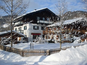 A multi-story building with a snow-covered roof, wooden balconies, and a wooden fence in a snowy landscape. - Holiday Ringberg - Gästehaus Schiffmann (Tegernsee)