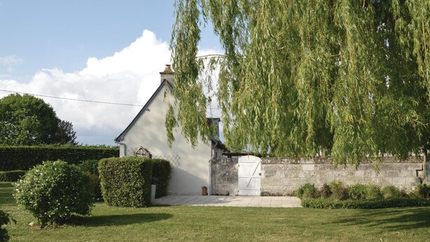 Un piccolo edificio bianco con un muro in pietra, una porta in legno e un salice piangente di fronte.