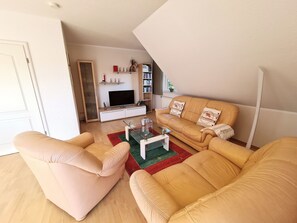 Un salon comprenant un canapé, des fauteuils, une table basse, un téléviseur et une bibliothèque.