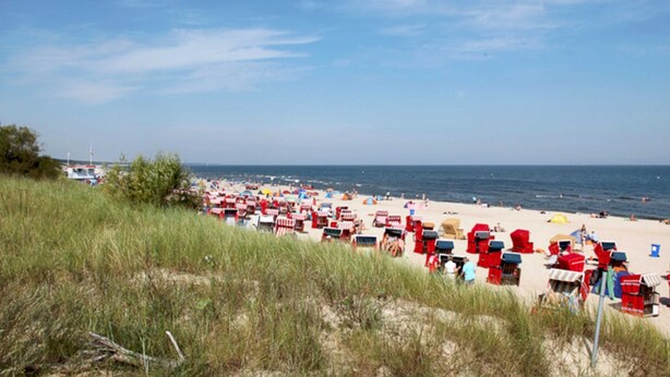 Ein Strand mit Reihen roter Liegen und Sonnenschirmen, ein sandiger Uferstreifen und das Meer im Hintergrund.