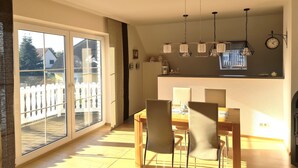 A dining area with a table and chairs, a kitchen with a clock and pendant lights, and a sliding glass door leading to a balcony.