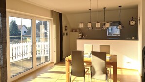 Ein Essbereich mit Tisch und Stühlen, eine Küche mit einer Uhr und Pendelleuchten sowie eine Glastür, die auf einen Balkon führt.