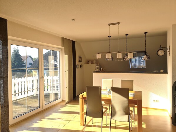 A dining area with a table and chairs, a kitchen with a clock and pendant lights, and a sliding glass door leading to a balcony.