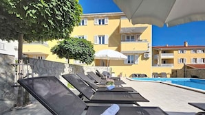 A poolside area with sun loungers, a pool, and a building with balconies in the background.