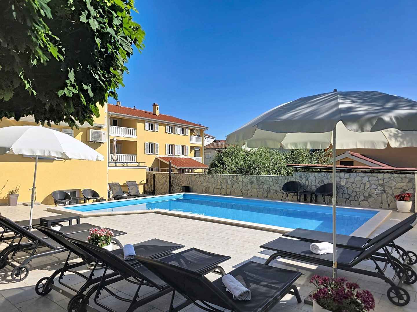 Una piscina con sillas de descanso y sombrillas, rodeada por un muro de piedra y edificios.