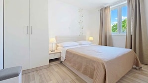 A bedroom with a bed, bedside tables, a wardrobe, a window with curtains, and a view of greenery outside.