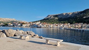 A waterfront promenade with benches, a clear view of a coastal town, and rocky terrain in the background.
