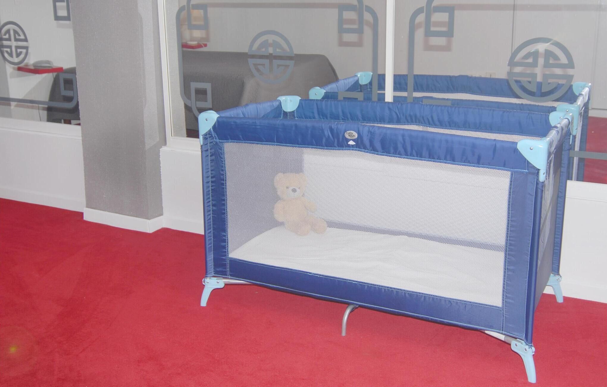 Un parc pour enfants bleu et blanc, avec un ours en peluche à l’intérieur, est placé sur un tapis rouge dans une pièce dotée d’une fenêtre et d’un mur blanc.