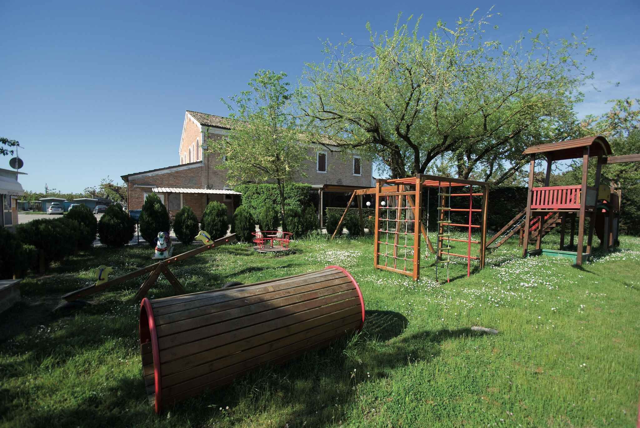 A playground with swings, a slide, and a climbing structure in a grassy area with trees and a house in the background.