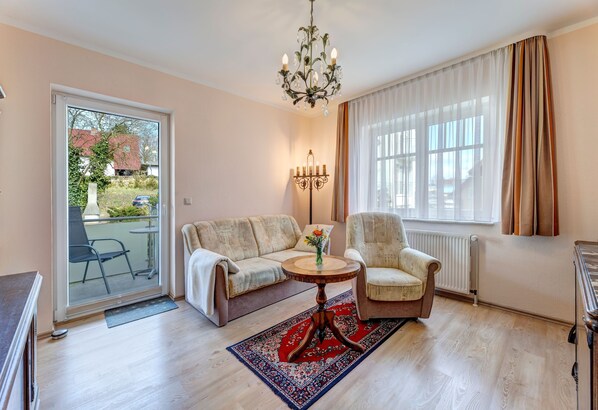 Un salon comprenant un canapé, un fauteuil, une table ronde, un lustre et une vue sur l’extérieur, accessible par une porte-fenêtre coulissante.