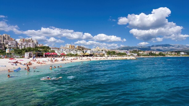 Une plage avec une eau d’un bleu limpide, des gens qui se baignent et des bâtiments en arrière-plan.