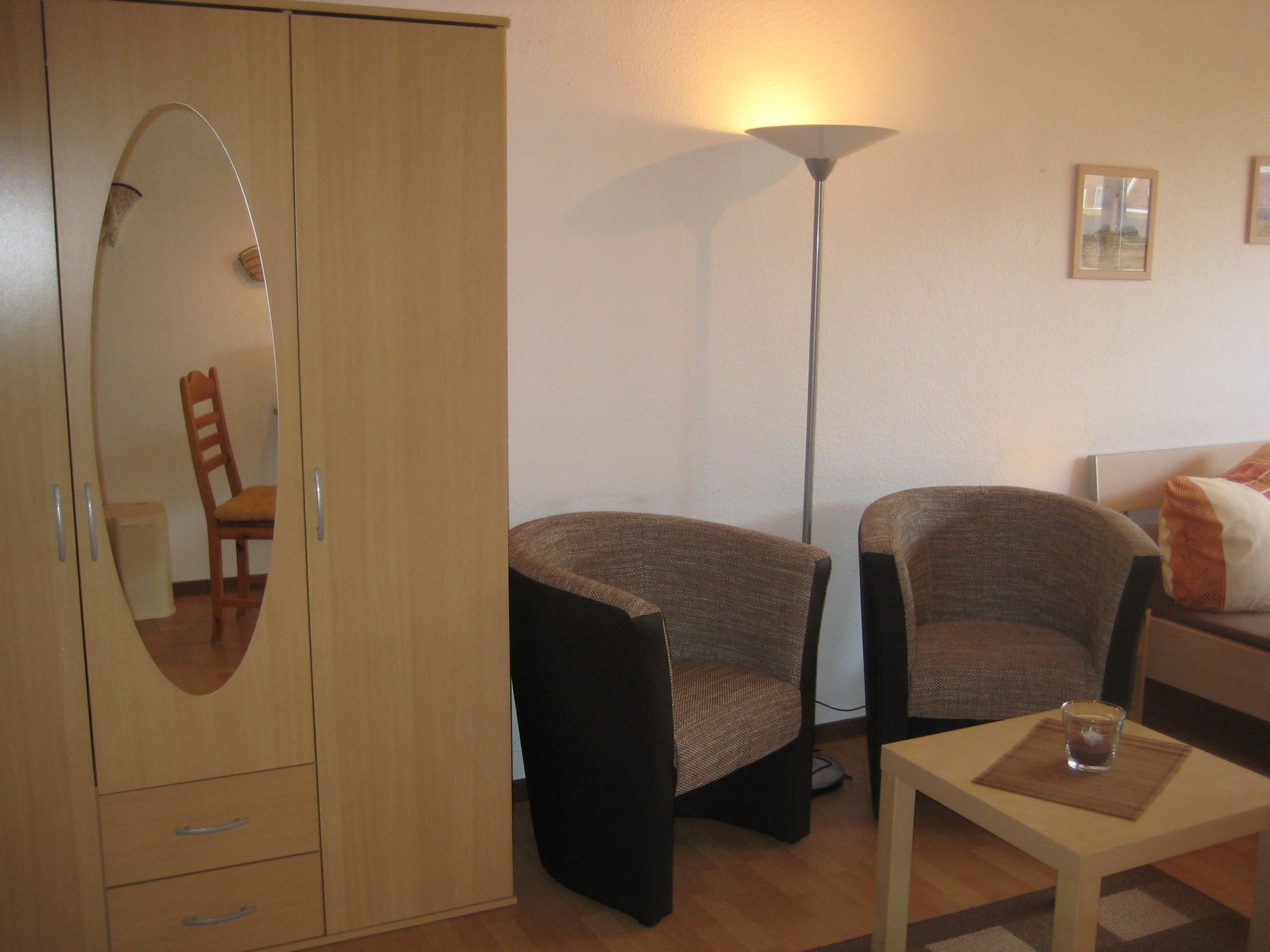 A room with a wooden wardrobe, two armchairs, a floor lamp, a small table with a glass, and a bed with pillows.