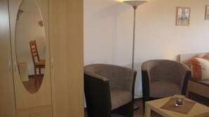 A room with a wooden wardrobe, two armchairs, a floor lamp, a small table with a glass, and a bed with pillows.