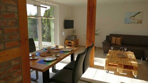 Un coin repas avec une table en bois, des chaises et un espace dédié au café. On aperçoit un canapé et un téléviseur au fond.