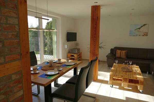 Un coin repas avec une table en bois, des chaises et un espace dédié au café. On aperçoit un canapé et un téléviseur au fond.
