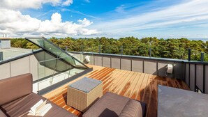 Een terras op het dak met een glazen balustrade, houten vloer en buitenmeubilair.