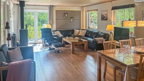Een woonkamer met een bank, een fauteuil, een salontafel en een eettafel met stoelen. Er zijn ramen met gordijnen, een radiator en een wandplank met decoratieve spullen.