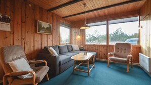 Een knus interieur met houten wanden, een grijze bank, twee gestreepte fauteuils, een klein houten tafeltje en een raam met uitzicht op bomen en een auto.