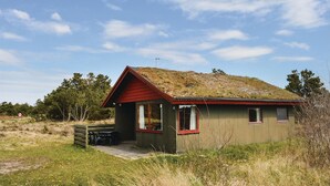 Een klein, gelijkvloers huisje met een rood dak en een groene gevel. Er is een klein terras met een tafel en stoelen, en een enkel raam aan de voorkant.