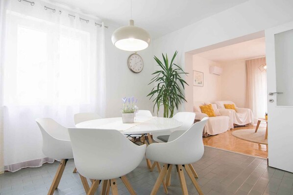 Un comedor moderno con una mesa redonda, sillas blancas, una planta en maceta y un gran ventanal con cortinas traslúcidas.
