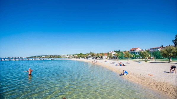 A beach with clear water, people sunbathing, and buildings in the background.