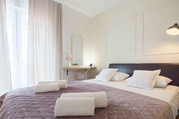 Una cama bien hecha con colcha marrón, almohadas blancas y dos toallas enrolladas. Hay una mesita de noche con un espejo y un pequeño jarrón con flores. La habitación tiene un diseño sencillo y limpio, con una pared de color claro y una lámpara de techo.