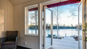 A room with a wicker chair, a wooden floor, and a large glass door leading to a wooden deck by a lake. - Stunning home in Holmsjö with kitchen (Holmsjö)