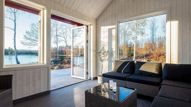 A modern interior with a sofa, a glass coffee table, and large windows offering views of a lake and trees. - Stunning home in Holmsjö with kitchen (Holmsjö)
