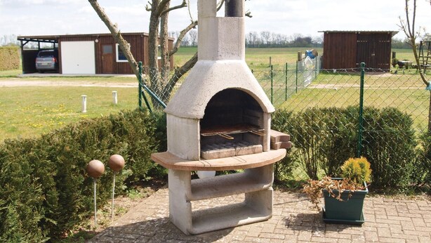 Un four à pizza extérieur en pierre, avec une base en briques, entouré d’une clôture en treillis métallique, situé dans un espace aménagé avec un bâtiment en bois en arrière-plan.